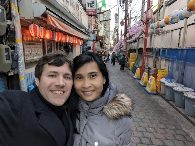 ggraph of Chris and Emily in a Japanese alleyway. There is a bar with lights behind them.